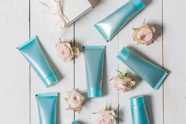 A few of the hand cream, primer, toner, face in blue, turquoise packing on a white wooden background with flowers of roses, overhead view — Stock Photo, Image