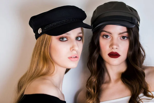 Deux belles femmes, copines, filles blondes et brunes en casquettes noires et maquillage lumineux regardent la caméra sur un fond blanc . — Photo