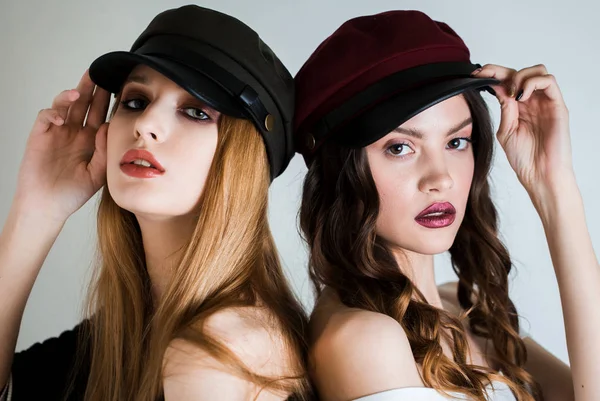 Deux femmes en casquettes et avec un maquillage lumineux sur fond blanc dos à dos regardant la caméra. Mode, cosmétique, soin du visage — Photo