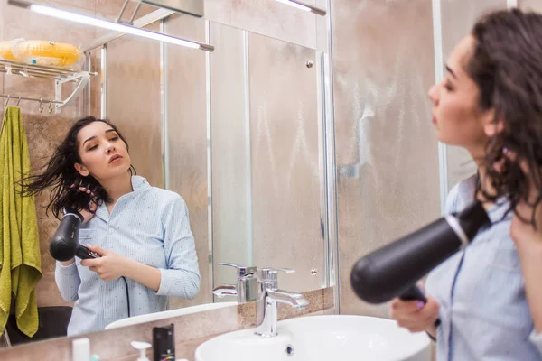 Wanita muda cantik dengan handuk mandi menggunakan pengering rambut dan melihat ke cermin — Stok Foto