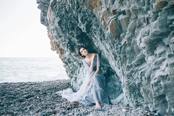 Belle fille brune en robe bleue assise près des rochers, mer, sourire , — Photo