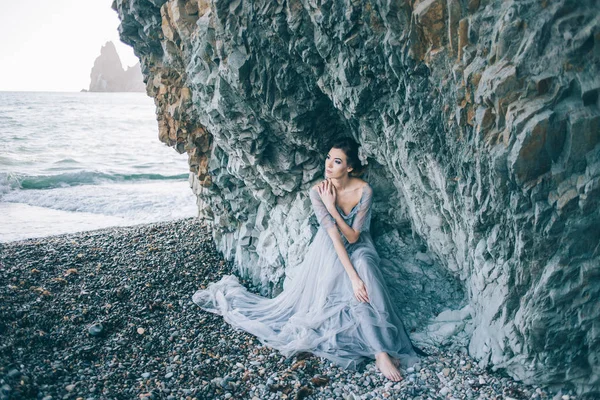 Krásná brunetka v dlouhé šedé šaty se závojem, sedí u moře, v blízkosti útesu, — Stock fotografie