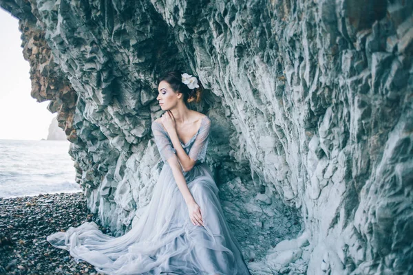 美丽的黑发女孩在蓝色礼服, 白色花在头发坐在附近悬崖, 海, — 图库照片