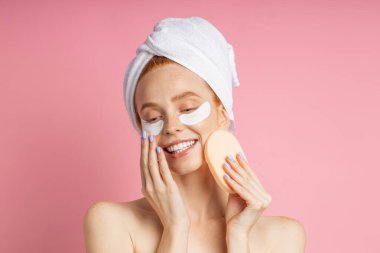 Genç bir kadının fotoğrafı göz bandı yapıştırıyor ve elinde kozmetik sünger tutuyor.