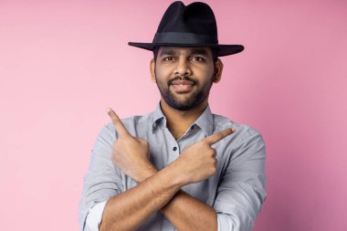 Tıraşsız, mutlu, genç, yakışıklı bir Hintli adamın, günlük gömlekli, şık siyah şapkalı, kameraya bakan ve işaret parmaklarıyla her iki yöne bakan, pembe stüdyo duvarına karşı duran.