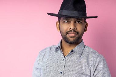 Küçük sakallı, gri gömlekli, siyah şapkalı, pembe arka planda poz veren, ciddi bir ifadeyle kameraya bakan yakışıklı Hintli genç adamın stüdyo portresi..