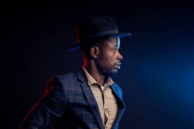 Stüdyoda genç, yakışıklı, kendine güvenen, ciddi, tıraşsız, Afrikalı iş adamı, gömlek giyen, kareli takım elbiseli, koyu arka planda duran, elinde siyah şapkayla duran,.