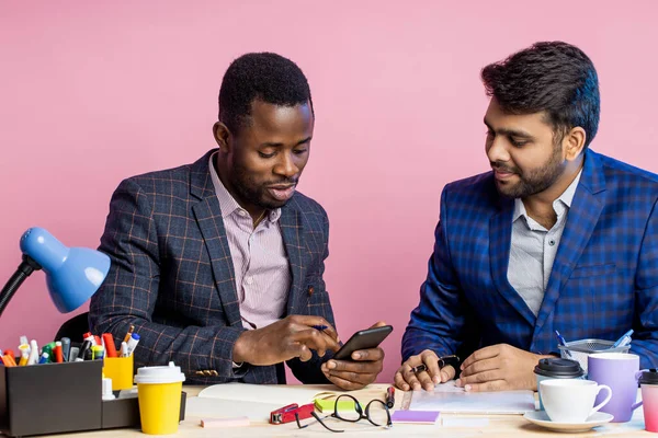 Two diverse business people in suits chatting, sitting at table in office. African young man showing his colleague new app on his phone, using mobile banking. Business, technology, work break concept