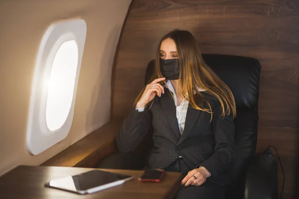 Jonge mooie zakenvrouw zittend in een vliegtuig met een zwart medisch masker tijdens een coronavirus pandemie — Stockfoto