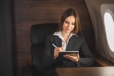 Çekici beyaz iş kadını uçakta rahat bir koltukta oturuyor, modern tableti tutuyor, gemideki kablosuz bağlantıyı kullanıyor. İş, seyahat ve teknoloji.