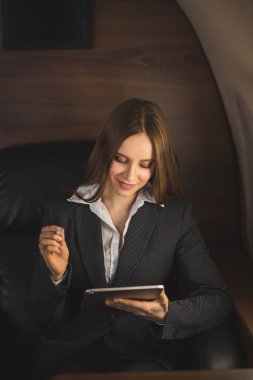 Siyah takım elbise giyen, tablet kullanan, özel iş uçağının koltuğunda otururken gülümseyen güzel, başarılı, kahverengi saçlı genç iş kadını. Birinci sınıf uçuyor..