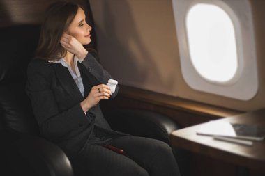 Kafkasyalı genç iş kadını rahat koltuğunda oturuyor, gülümsüyor, elinde modern kablosuz kulaklıklar tutuyor, uzun uçuşlar boyunca sesli kitap dinleyecek. Teknoloji konsepti.