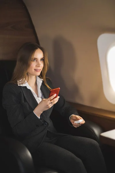 Beautiful young businesswoman traveling in private jet. Brown haired lady in white blouse, formal suit holding smartphone, sitting in comfortable seat in airplane.