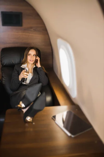 Confident self assured young businesswoman sitting in private plane with her feet on table, talking on mobile phone, holding glass of champagne, wearing formal black suit, high heel shoes.