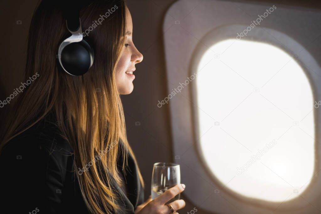Closeup shot of happy young caucasian woman with long blonde hair, in black jacket dress, listening to music with headphones, holding glass of champagne, looking at airplane window with cute smile.