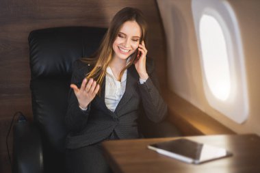 Neşeli kahverengi saçlı, beyaz bluzlu, resmi takım elbiseli, rahat koltukta oturan, birinci sınıf uçakla uçarken cep telefonuyla konuşan neşeli iş kadını..
