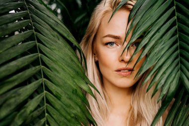 Genç bir kadının yüzü tropik yapraklarla çevrili. Doğal çıplak makyajlı, mükemmel ciltli, sarı saçlı muhteşem bir kadının yakın plan portresi. Doğal kozmetik, sağlık, saflık, cilt bakımı, güzellik konsepti.
