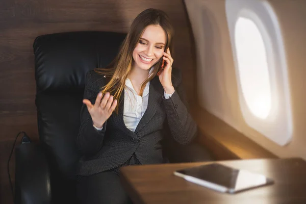 Cheerful happy brown haired business lady in white blouse and formal suit, sitting in comfortable seat, laughing talking on mobile phone during flight on first class plane.