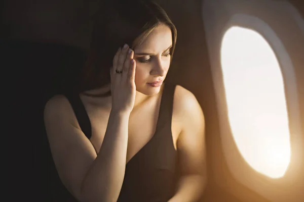 Closeup shot of beautiful tired girl sitting in airplane at window, touching her head, experiencing stress, headache, migraine. Young woman is afraid to fly. Fear of flying, pain, aerophobia concept.
