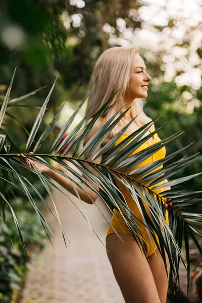 温和的女领事 金发碧眼 身材苗条 身穿黄色泳衣 日落时在热带植物的背景下摆姿势 夏天的概念 镜头聚焦在树叶上 — 图库照片