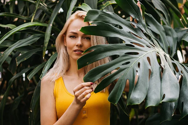 迷人的女孩在热带雨林里摆姿势 年轻的高加索女人的肖像 金发碧眼 美丽的眼睛从热带叶子向外看相机 夏季概念 — 图库照片