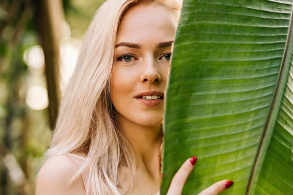 Sarışın, doğal makyajlı, temiz tenli, yüzünün yarısını tropikal yaprağın arkasına saklayan beyaz bir kadının yakın plan portresi. Cilt bakımı, kaplıca, güzellik, organik kozmetik ürünler konsepti.