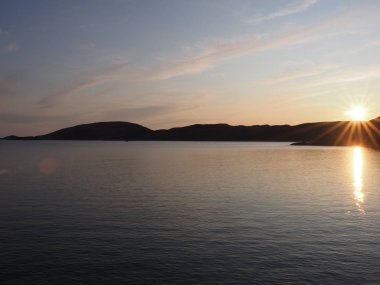 Norveç 'in Salten bölgesindeki Avrupa Bodo şehrindeki suya yansıyan güneş batışına, 2019 yılında güneşli bir yaz akşamında açık mavi gökyüzüne yansıyor..