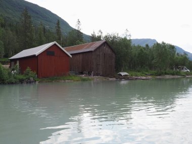 Norveç 'in Nordland ilçesinde Svartisvatnet Gölü' ndeki ahşap barakalar ve dağlar Avrupa 'nın Svartisen buzulu yakınlarındaki suya, 2019 yılının soğuk yaz gününde bulutlu gökyüzüne yansıdı..