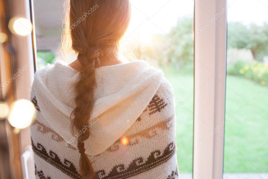 Thoughtful young brunette woman wearing nordic print poncho looking through the window, blurry garden outside
