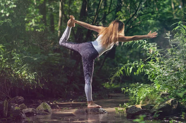 Woman in the woods standing on the creek rock practicing yoga, wellness and wellbeing concept.