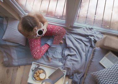 Young beautiful sitting down on the floor by the window with cup of coffee, winter christmas home concept, snow outside