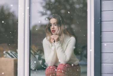 Snowflakes by the window. Young beautiful blonde woman with red lips sitting home in living room. Winter snowing concept
