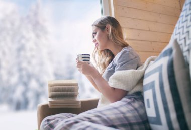 Young beautiful blonde woman with cup of coffee sitting home in living room by the window. Winter snow landscape view. Christmas concept