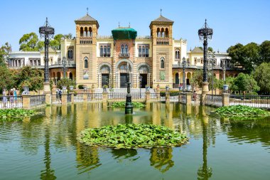 Seville, İspanya - Maria Louise Parkındaki Arkeoloji Müzesi Pavyonu, bej renkli bir bina, ön planda yeşil zambak yapraklı bir gölet, mavi gökyüzü, yaz öğleden sonra.