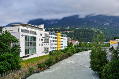 Landeck, Austria - 06.26.2014: White rectangular buildings in a small town on the banks of a mountain river among green trees, amid alpine mountains covered with clouds, in the summer afternoon.