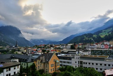 Landeck, Avusturya - Küçük bir kasabadaki beyaz evlerin yanında turuncu bir ev, ormanla kaplı yeşil dağların arasında, gökyüzünde gri bulutlar, yaz öğleden sonra.