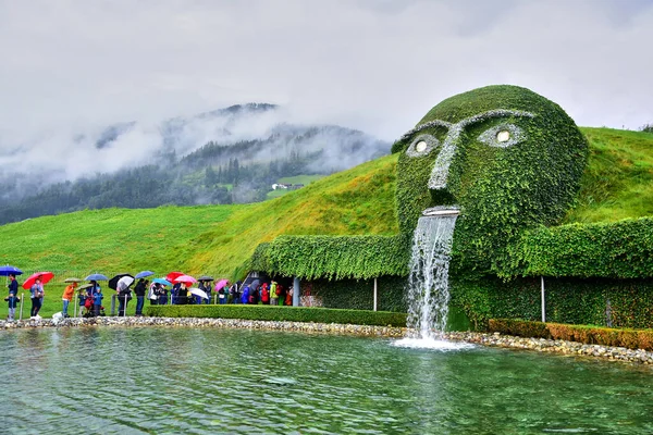 Wattens, Avusturya - 08.09.2916: Swarovski Kristal Müzesi, bitkilerle kaplı bir çeşme, ağızdan su akıyor, şemsiyeli insanlar, yeşil tepeler, bulutlar.