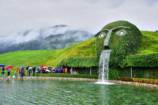 Wattens, Austria - 08.09.2916: Swarovski Crystal Museum, a fountain in the form of a head covered with plants, water flows from the mouth, people with umbrellas, green hills, clouds.