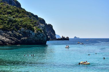Paleokastritsa / Korfu, Yunanistan - Teknelerin ve insanların yelken açtığı turkuaz bir deniz, yeşil ağaçlı ve çalılıklı gri kayalar, yaz öğleden sonra mavi bir gökyüzü.
