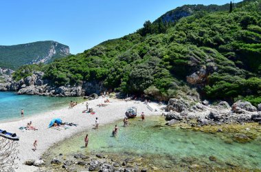 Paleokastritsa / Korfu, Yunanistan - Limni Sahili, beyaz çakıl taşı plajı, sahilin her iki tarafında su, arka planda yeşil ağaçlarla kaplı bir dağ, kıyıda dinlenen turistler.