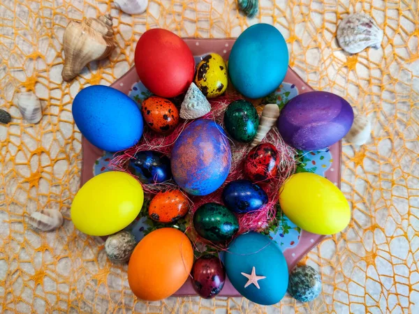 Religious Easter holiday, on an orange grid lies a plate with multi-colored chicken and quail eggs, sea shells and shells are laid around them.