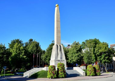 Cesis, Letonya - 07.22.2017: Cesis 'te Özgürlük Anıtı, üzerinde altın bir top bulunan yüksek bir taş dikilitaş, mor çiçekler, ağaçlar, mavi gökyüzü bulunan yeşil çiçek yataklarının yanında, yazın.