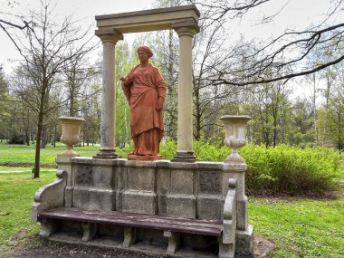 Frantiskovy Lazne, Çek Cumhuriyeti - 05.03.2016: Yolun kenarındaki bir parkta, bir bankın yanında, çiçek vazoları, yeşil çalılar ve ağaçlı taş kolonlar olan bir heykel, gündüz vakti.