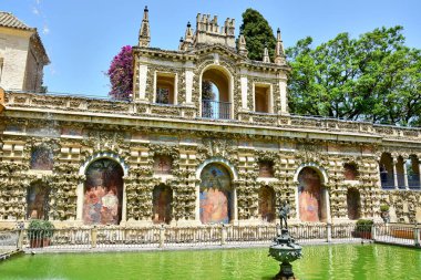 Sevilla, İspanya - 07.21.2019: Seville Alcazar, aslen Mağripliler 'in kalesi, süs ve resimlerle dolu bir duvar, yeşil suyu, çeşmesi ve heykeli olan yapay bir gölet, mavi bir gökyüzü.