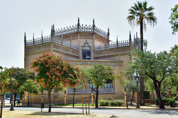Seville, İspanya - 07.21.2019: Pabellon Real, bir belediye kurumu, dekorla süslenmiş kahverengi bir bina, ön plandaki ağaçlar ve bir palmiye ağacı, ağaçtaki kahverengi çiçekler.
