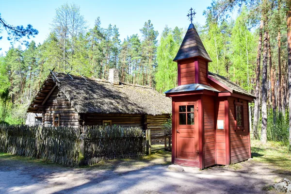 Riga, Letonya - Letonya Açık Hava Müzesi, küçük bir bordo ahşap şapel ve sazdan çatılı ve çitli gri bir köy evi, arka planda yeşil ağaçlar.