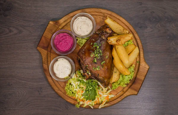 Fried pork leg drumstick, knee of a wild boar with vegetables of red and yellow tomatoes, garlic, parsley and basil and potatoes on a wooden background — Stock Photo, Image