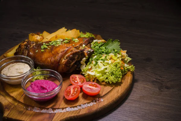 Fried pork leg drumstick, knee of a wild boar with vegetables of red and yellow tomatoes, garlic, parsley and basil and potatoes on a wooden background — Stock Photo, Image