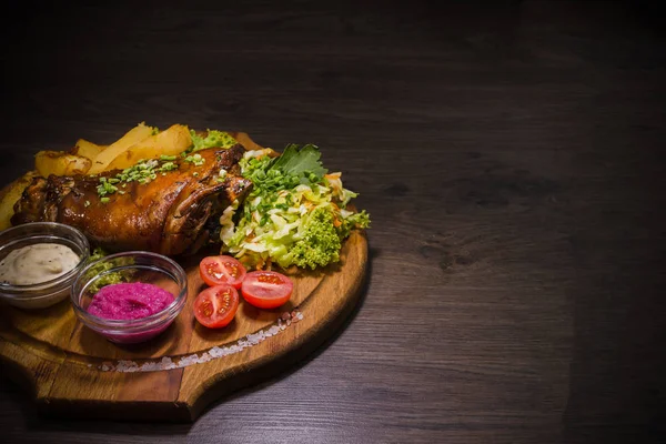 Fried pork leg drumstick, knee of a wild boar with vegetables of red and yellow tomatoes, garlic, parsley and basil and potatoes on a wooden background — Stock Photo, Image