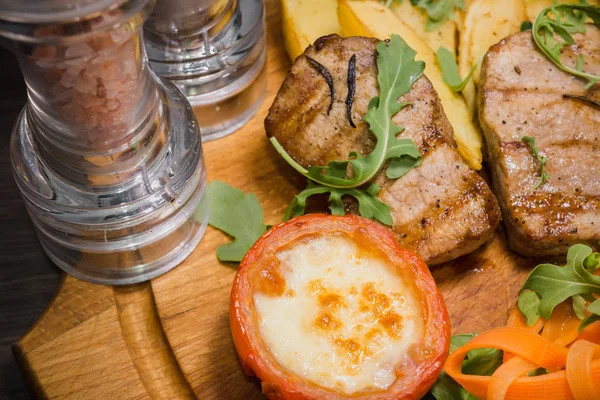 Three pork steaks with fried potatoes and vegetables on a wooden background — Stock Photo, Image